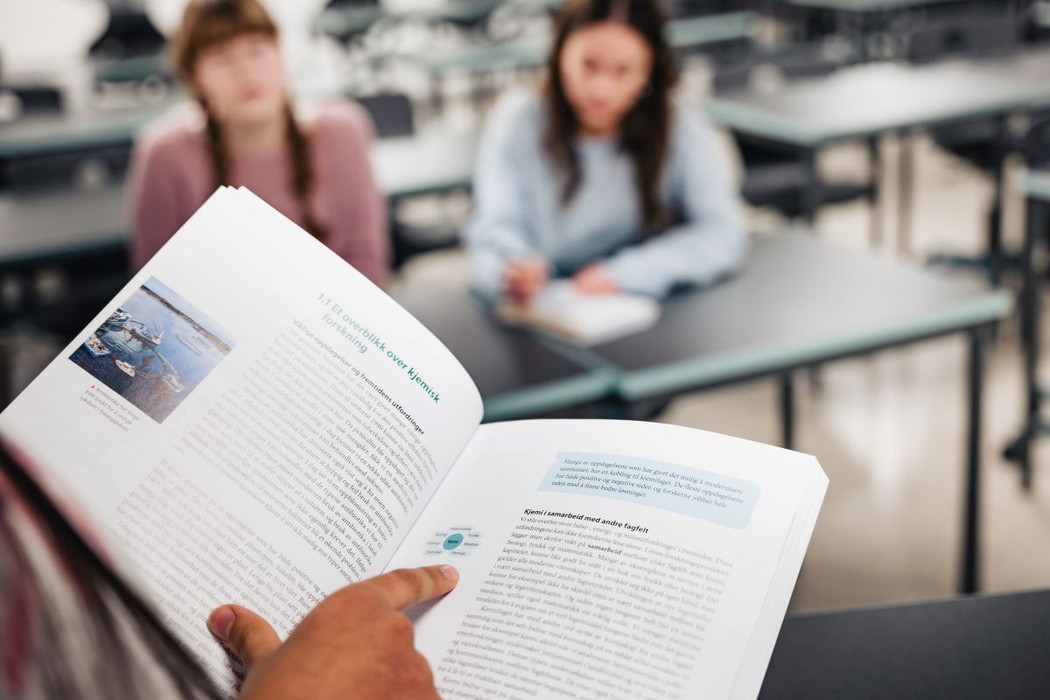 Oppslått lærebok i fokus holdt av kvinne som peker på figur. I bakgrunn sitter to ufokuserte elever.