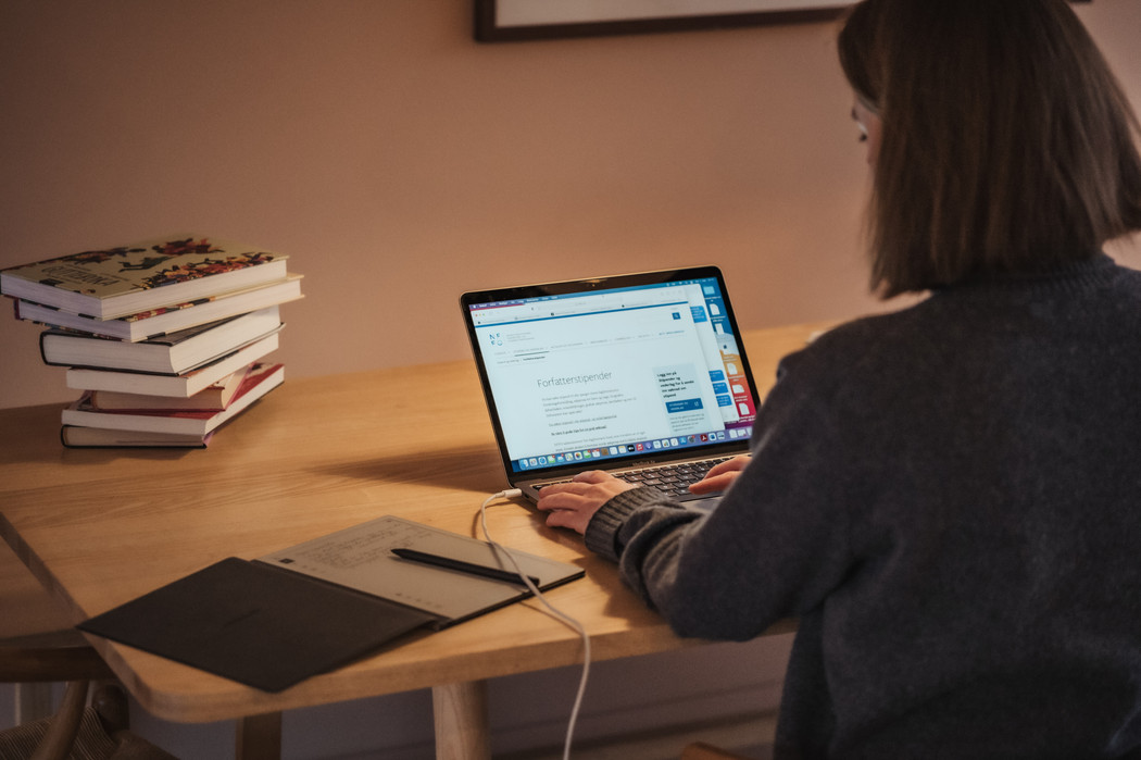 Kvinne sitter med ryggen til ved et spisebord. Hun skriver på en laptop som viser stipendsidene på nffo.no. På bordet ligger også en e-notatblokk og en bunke med bøker. 