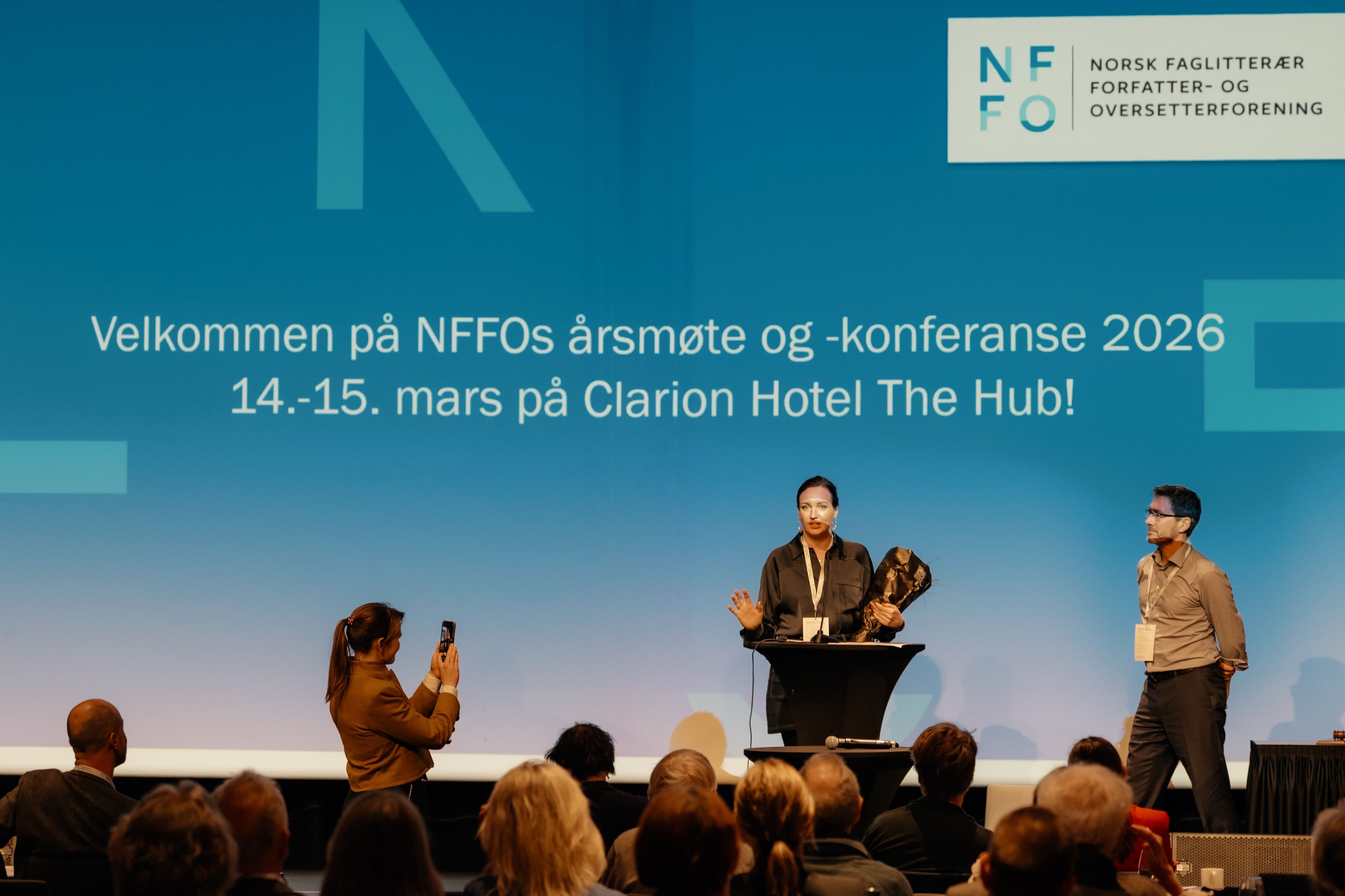 Kvinne på scene foran bakteppe hvor det står "Velkommen på NFFOs årsmøte og -konferanse 2026 14.-15. mars på Clarion Hotel The Hub!"