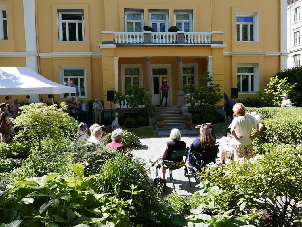 Oversiktsbilde av gul villa i nyklassisk stil med veranda med søyler. På verandaen en mann som holder tale. I forgrunnen en frodig hage med mennesker som står og sitter. 