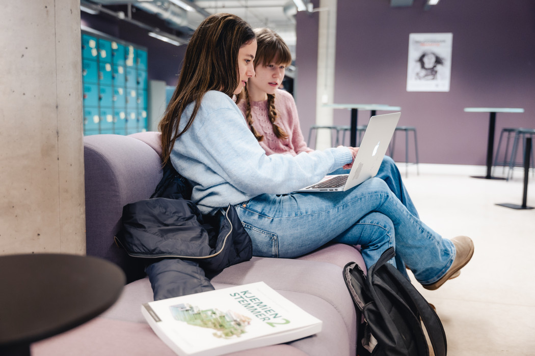 To tenåringsjenter sittere i en sofa i et vrimleormåde på en skole. Den ene har en laptop på fanget som de begge ser på.