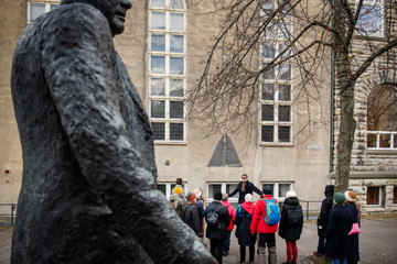 I forgrunnen deler av en skulptur av en mann, i bakgrunnen en gruppe mennesker som er på byvandring, midt i gruppen står en mann med utstrakte armer og snakker. 