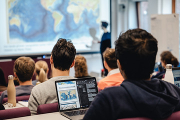 Studenter i et auditorium sett bakfra. I bakgrunnen en foreleser foran et lerret som viser et verdenskart.