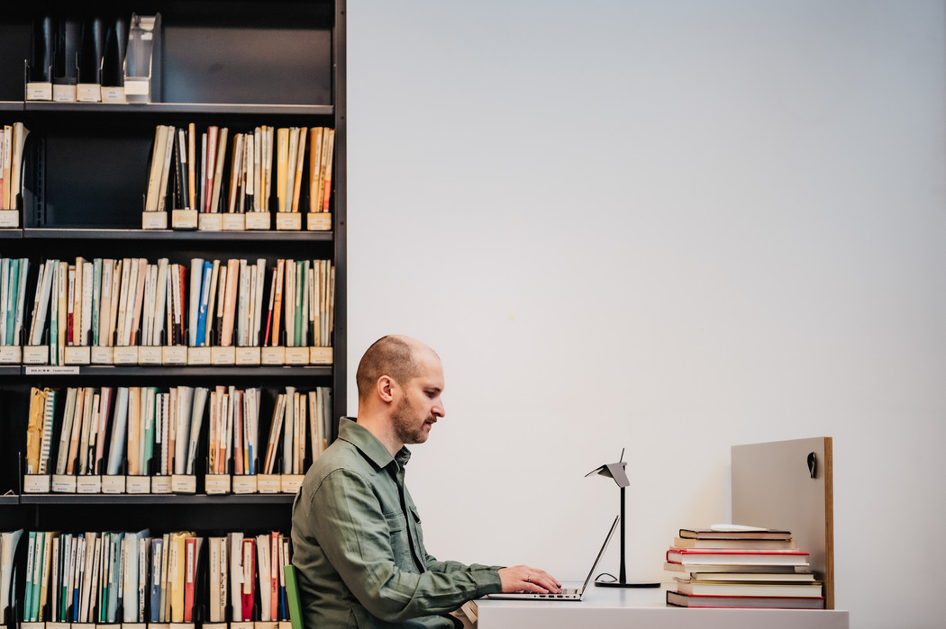 Mann som sitter ved en pult på et bibliotek og skriver på en laptop. På pulten er det en lampe og en stabel med bøker. I bakgrunnen er det en bokhylle med permer og mapper.