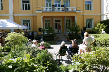Oversiktsbilde av gul villa i nyklassisk stil med veranda med søyler. På verandaen en mann som holder tale. I forgrunnen en frodig hage med mennesker som står og sitter. 
