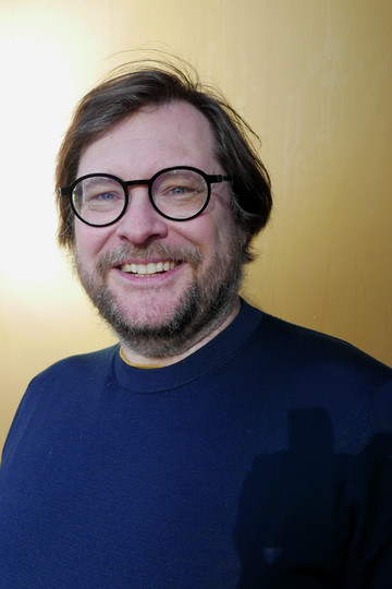Mann med briller, skjegg og blå genser foran en gullfarget bakgrunn.